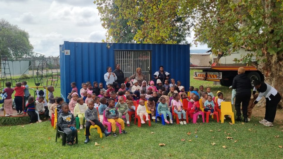 A group of young children seated outside a blue container classroom with caregivers, highlighting early childhood learning in a community setting.
