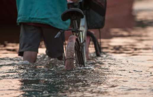 Person walking a bicycle through floodwater in a flooded community street during severe weather conditions.