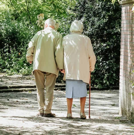An elderly couple walking together outdoors along a garden pathway.