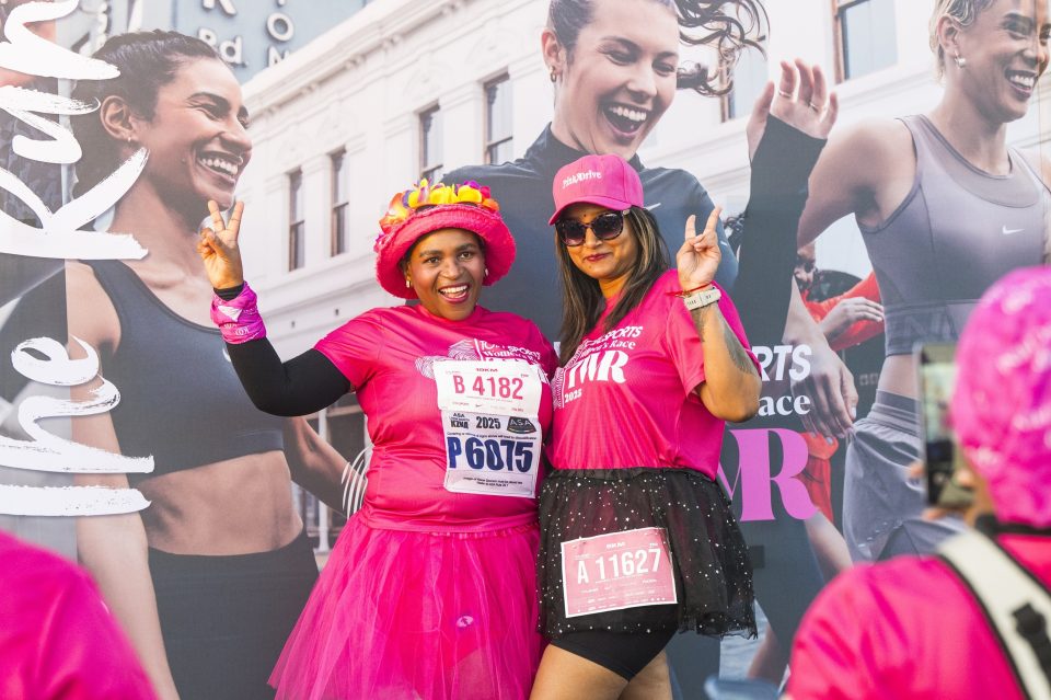 Two women in pink outfits posing at the Totalsports Women’s Race, celebrating breast cancer awareness with race numbers and PinkDrive branding visible.