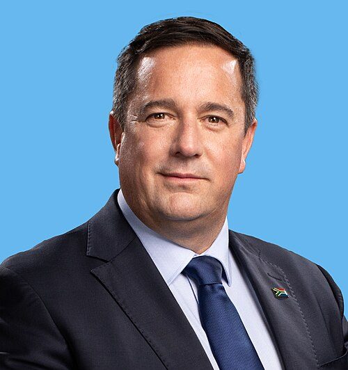 South African Minister of Agriculture John Steenhuisen during an official portrait session, wearing a dark suit and tie against a blue background.