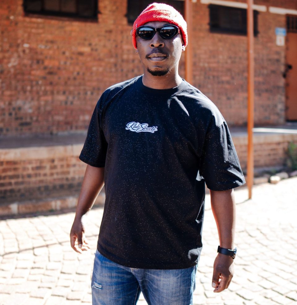 Content creator and cultural commentator Madala TK standing outdoors wearing a black T-shirt, red beanie and sunglasses.