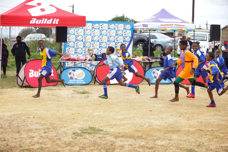 Young footballers sprint across a dusty community football pitch during a Build it U13 Football Tournament match, cheered on by teammates, coaches and spectators.