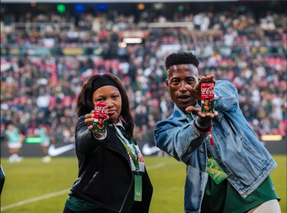 Two rugby fans hold limited-edition Coca-Cola Springbok Fan Cans while standing pitch-side at a packed rugby stadium.