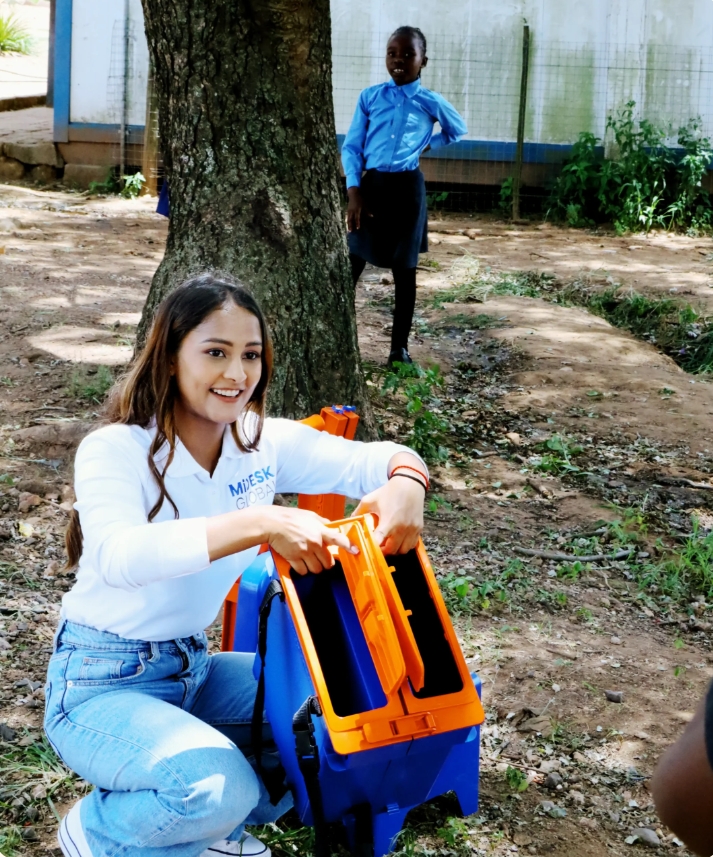 A MiDesk Global volunteer demonstrates a portable school desk during a school visit while a learner watches nearby, highlighting efforts to improve classroom learning conditions.