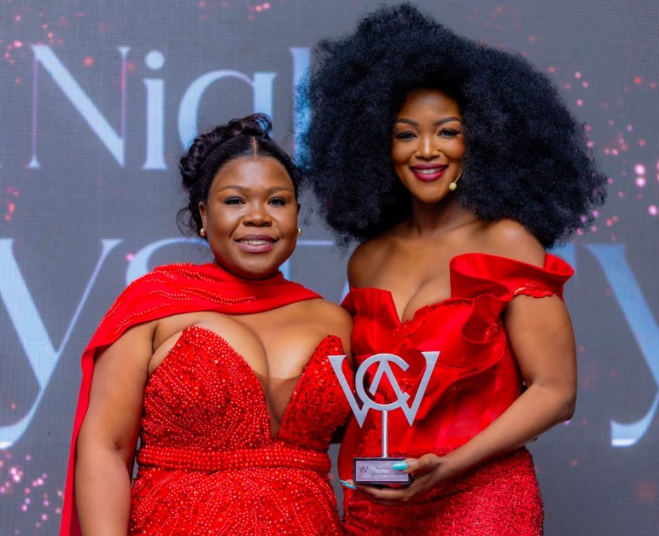 Two women dressed in red at the WomenCan Awards, with one holding a WomenCan award on stage.