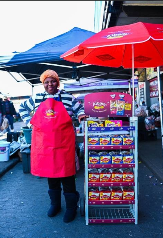 Albany Bread-On-The-Streets entrepreneur standing beside a branded bread stand stocked with Albany products.