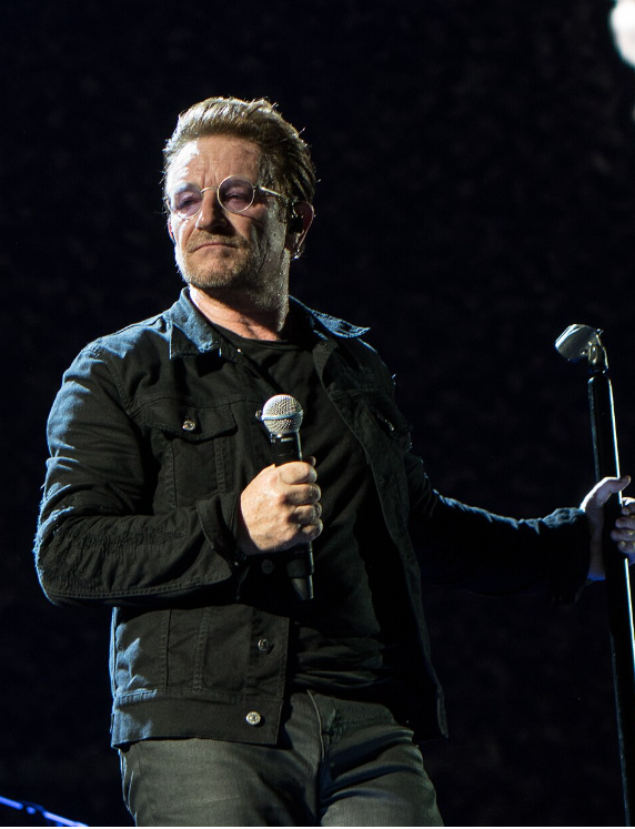 Bono performing on stage with U2, holding a microphone during a live concert.