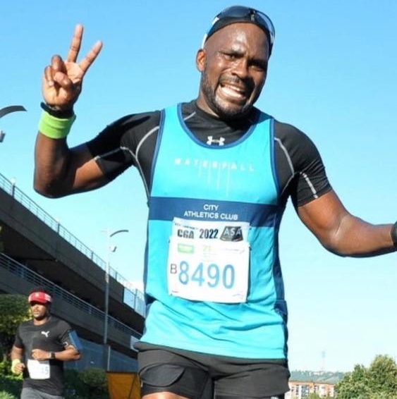 Endurance athlete Temo Jacopo running in a road race, raising a peace sign while wearing a blue athletics vest.