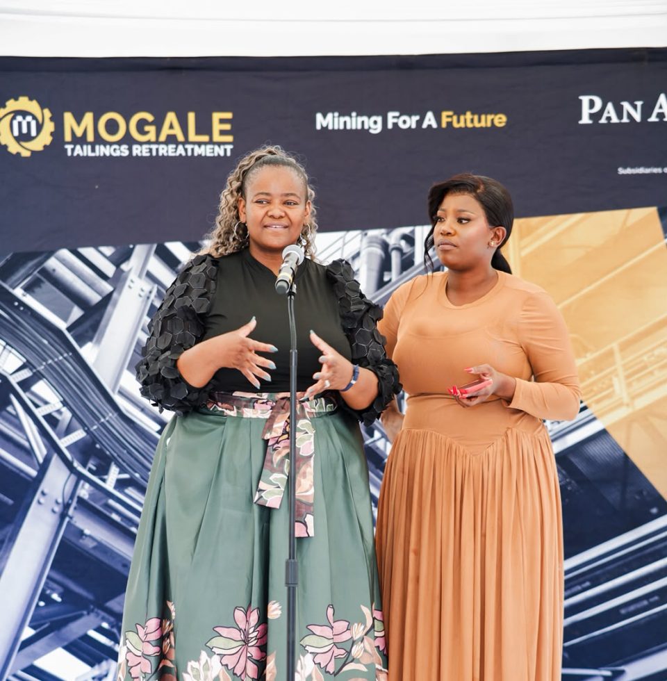 Two women address attendees at a Mogale Tailings Retreatment community education graduation ceremony, standing together at a microphone.