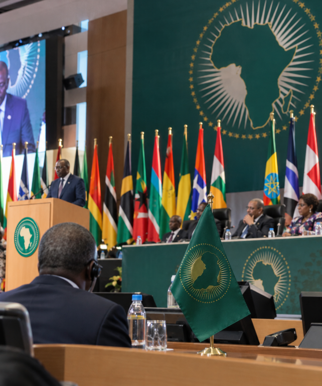 Delegates attend an African Union-style conference as a speaker addresses the room from a podium, with multiple African flags and a continental emblem visible.